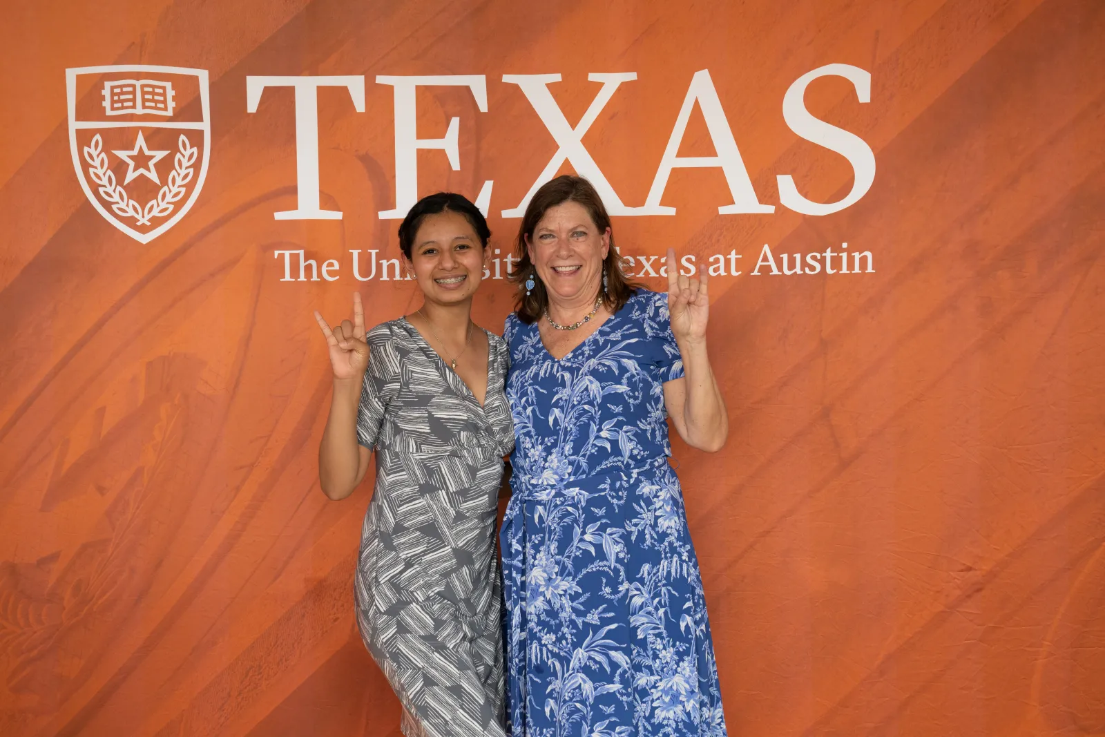 mentor and mentee smiling holding up the hookem horns handsign in front of a burnt orange background with the UT Austin wordmark on it.