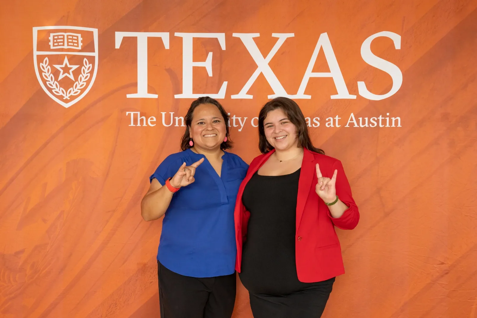 a staff mentor and mentee posing together doing the HookEm Horns Handsign in front of a burnt orange background with "TEXAS The University of Texas at Austin" logo.
