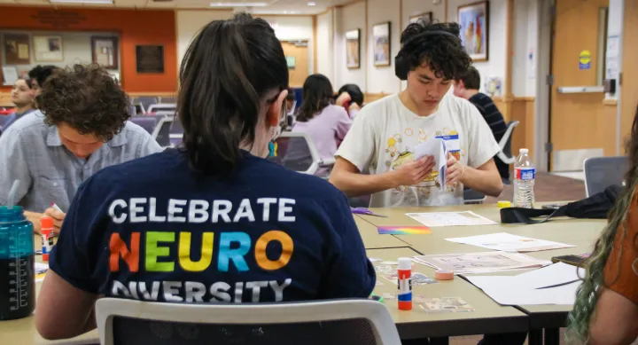 Two students crafting at a table next to a student worker wearing a shirt that says "Celebrate Neuro Diversity" 