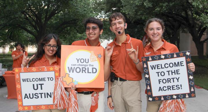4 students in orange polos holding signs that say "Welcome to UT Austin". The Students are also holding pom poms.