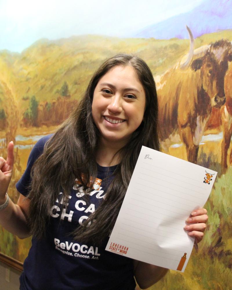student holding up a piece of paper with lines on it and doing a hookem with the other hand